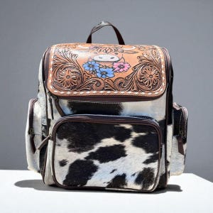 May include: A cowhide backpack with a brown leather flap featuring a carved floral design and a cartoon cow with blue and pink flowers. The backpack has a top handle, multiple pockets, and a black and white cowhide pattern.