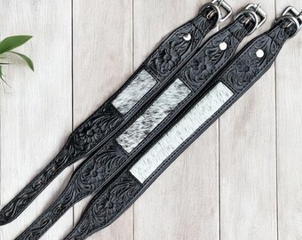 May include: Three black leather guitar straps with floral tooling and silver hardware. Each strap features a rectangular panel of cowhide with a black and white pattern. The straps are arranged diagonally on a light wood surface.