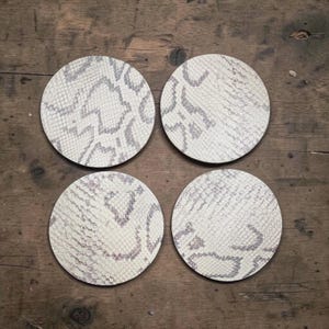 May include: Four round coasters with a snakeskin pattern in shades of cream and gray. The coasters are arranged on a wooden surface, offering a neutral and stylish accent for any home decor. These coasters are designed to protect surfaces from drink rings.