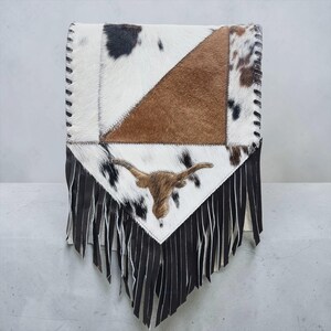 May include: A cowhide leather bag with a patchwork design in white, brown, and black. The bag features a longhorn emblem and black fringe along the bottom edge. The stitching is black.