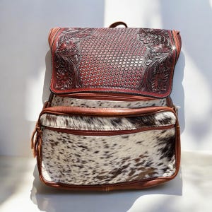 May include: A brown leather backpack with a decorative floral design on the flap. The lower section features a cowhide pattern in white and brown. The bag has a top handle and two zippered compartments.