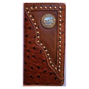 May include: Brown leather wallet with a textured ostrich pattern and gold-colored stud accents. Features a central brown leather panel and a decorative metal emblem with a cross and a landscape design. A stylish accessory.