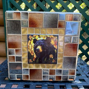 Könnte beinhalten: Quadratisches Mosaikkunstwerk mit einem Porträt eines schwarzen Bären in der Mitte, umgeben von braunen, goldenen und silbernen Fliesen. Der Bär hat ein detailliertes Gesicht und florale Akzente, was ein rustikales, dekoratives Objekt schafft.