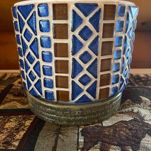 May include: A mosaic-style planter with a geometric pattern of blue and brown tiles. The planter has a white rim and sits on a round, textured base. The design is a mix of squares and diamond shapes.