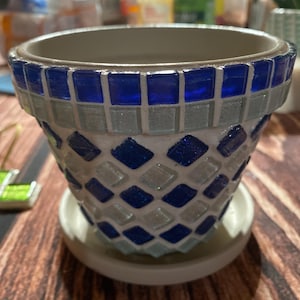 May include: A mosaic-tiled ceramic flower pot with a matching saucer. The pot features a pattern of blue and light blue tiles. The rim of the pot is lined with a row of blue tiles. The pot is designed for indoor or outdoor use.