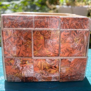 May include: A square decorative planter with a mosaic design. The planter is made of small, square tiles with a leaf pattern in shades of orange, brown, and yellow. The planter has a light-colored rim and is sitting on a blue surface.