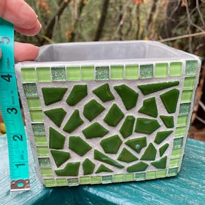 May include: A square, gray planter with a mosaic design. The planter is decorated with green glass tiles in a mix of square and irregular shapes. The border features small, square tiles and glittery accents. The planter measures approximately 4 inches in height.