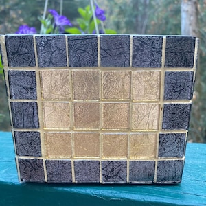 May include: Square mosaic planter with alternating black and gold tiles. The planter has a rectangular shape and is made of small, square tiles. The gold tiles are in the center, and the black tiles are around the edges. The planter is sitting on a green surface.