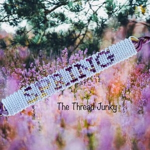 May include: White friendship bracelet with the word "SCORPIO" woven in purple and gold thread. The bracelet is photographed against a background of purple flowers.