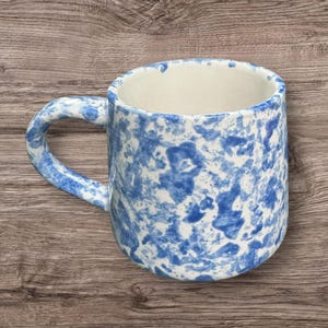 May include: A ceramic mug with a blue and white speckled pattern. The mug has a rounded body, a white interior, and a curved handle. The mug is sitting on a wooden surface.