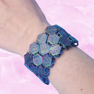 May include: A watch band composed of interconnected, iridescent hexagonal links that display a shifting spectrum of blue, purple, and green hues. The structure features a flexible, articulated design with a secure clasp mechanism at the end.
