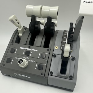May include: A gray Thrustmaster Boeing 737 flight simulator control panel with two throttle levers, a flap lever, and a variety of buttons and switches. The panel features a white knob labeled "SEL" and a white label that reads "FLAP".