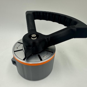 May include: A gray and orange mechanical device with a black handle. The handle is arched and textured for grip. The device has a circular base with the words "RIGHT" and "LEFT" visible.