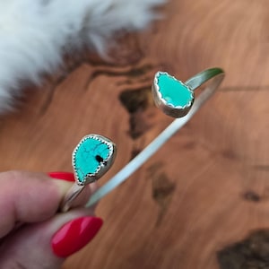 May include: Open cuff bracelet with turquoise teardrop-shaped stones set in silver. The turquoise stones have a vibrant teal color and are surrounded by a silver border. The bracelet is displayed on a wooden surface.