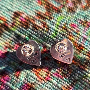 May include: A pair of heart-shaped sterling silver stud earrings. Each earring features a sparkling, round-cut diamond at the center. The word "STERLING" is engraved on each earring, along with a small heart. The earrings are displayed on a colorful, textured surface.