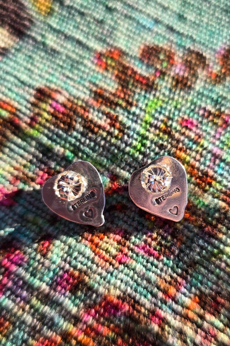 May include: A pair of heart-shaped sterling silver stud earrings. Each earring features a sparkling, round-cut diamond at the center. The word "STERLING" is engraved on each earring, along with a small heart. The earrings are displayed on a colorful, textured surface.