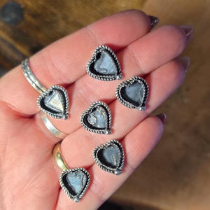 May include: Heart-shaped stud earrings with a silver-colored rope-like border and a light blue center. The earrings are displayed on a hand, showcasing their delicate design and craftsmanship. A ring is also visible.