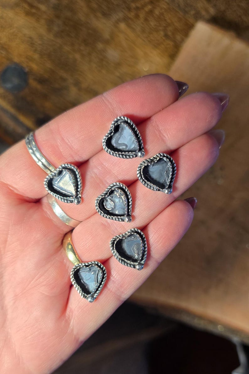 May include: Heart-shaped stud earrings with a silver-colored rope-like border and a light blue center. The earrings are displayed on a hand, showcasing their delicate design and craftsmanship. A ring is also visible.