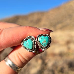 May include: A pair of heart-shaped stud earrings with turquoise centers and silver-toned rope and sawtooth borders. The earrings are displayed against a blurred natural background.
