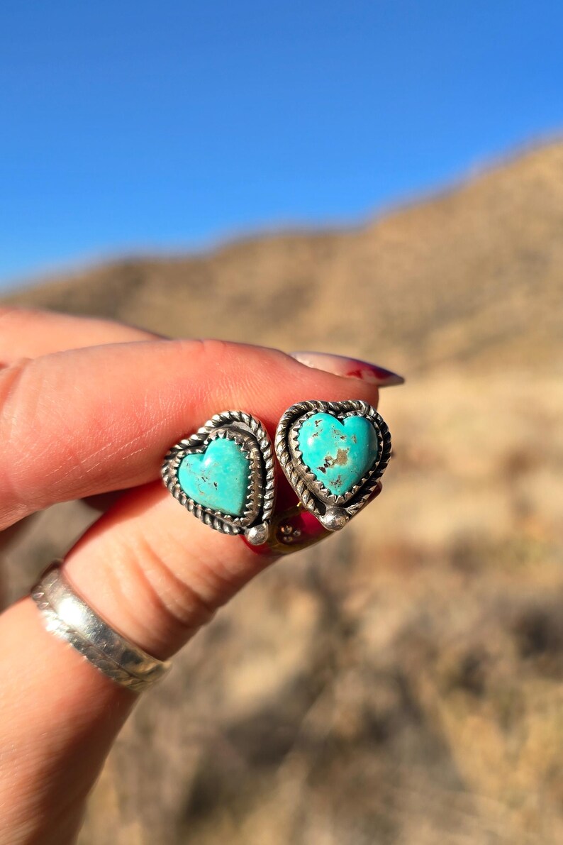 May include: A pair of heart-shaped stud earrings with turquoise centers and silver-toned rope and sawtooth borders. The earrings are displayed against a blurred natural background.