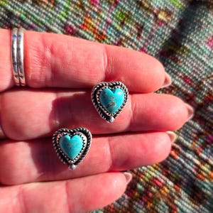 May include: A pair of heart-shaped stud earrings. Each earring features a turquoise stone set in a silver-toned frame with a twisted rope design. The earrings are displayed on a hand.