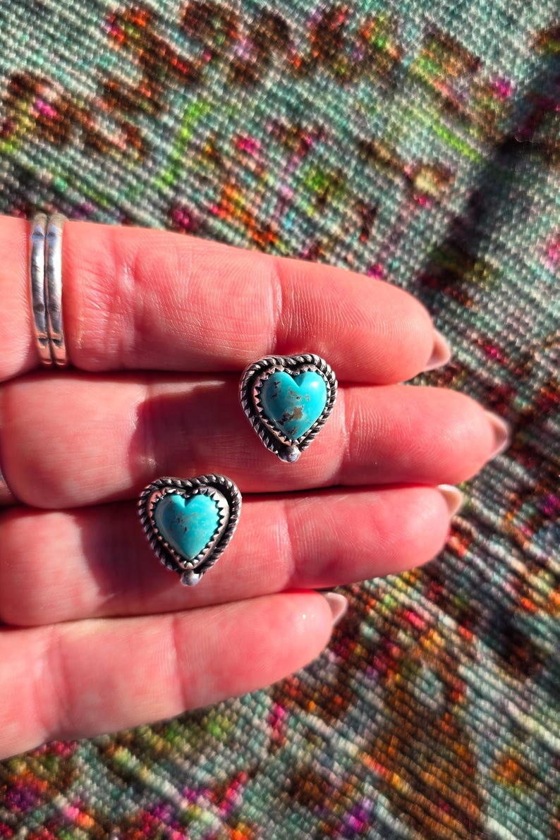 May include: A pair of heart-shaped stud earrings. Each earring features a turquoise stone set in a silver-toned frame with a twisted rope design. The earrings are displayed on a hand.