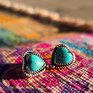 May include: A pair of heart-shaped stud earrings with turquoise centers and silver-colored, rope-textured borders. The earrings are displayed on a colorful, patterned textile with shades of pink, purple, orange, and yellow.