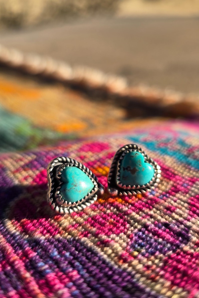 May include: A pair of heart-shaped stud earrings with turquoise centers and silver-colored, rope-textured borders. The earrings are displayed on a colorful, patterned textile with shades of pink, purple, orange, and yellow.