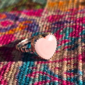 Puede incluir: Un anillo de plata con una piedra en forma de corazón de color rosa pálido. El anillo tiene una banda texturizada y un borde festoneado alrededor del corazón. Se encuentra sobre una tela colorida estampada.