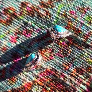 May include: A pair of silver stud earrings with teardrop-shaped turquoise stones. The earrings have a detailed, textured silver border. The background is a colorful, patterned textile with hues of pink, green, and brown.