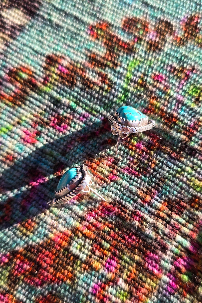 May include: A pair of silver stud earrings with teardrop-shaped turquoise stones. The earrings have a detailed, textured silver border. The background is a colorful, patterned textile with hues of pink, green, and brown.