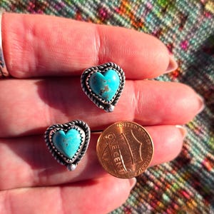 May include: A pair of heart-shaped turquoise stud earrings with a silver rope-like border. A U.S. one-cent coin is next to the earrings for size comparison. The earrings are set against a colorful, patterned background.