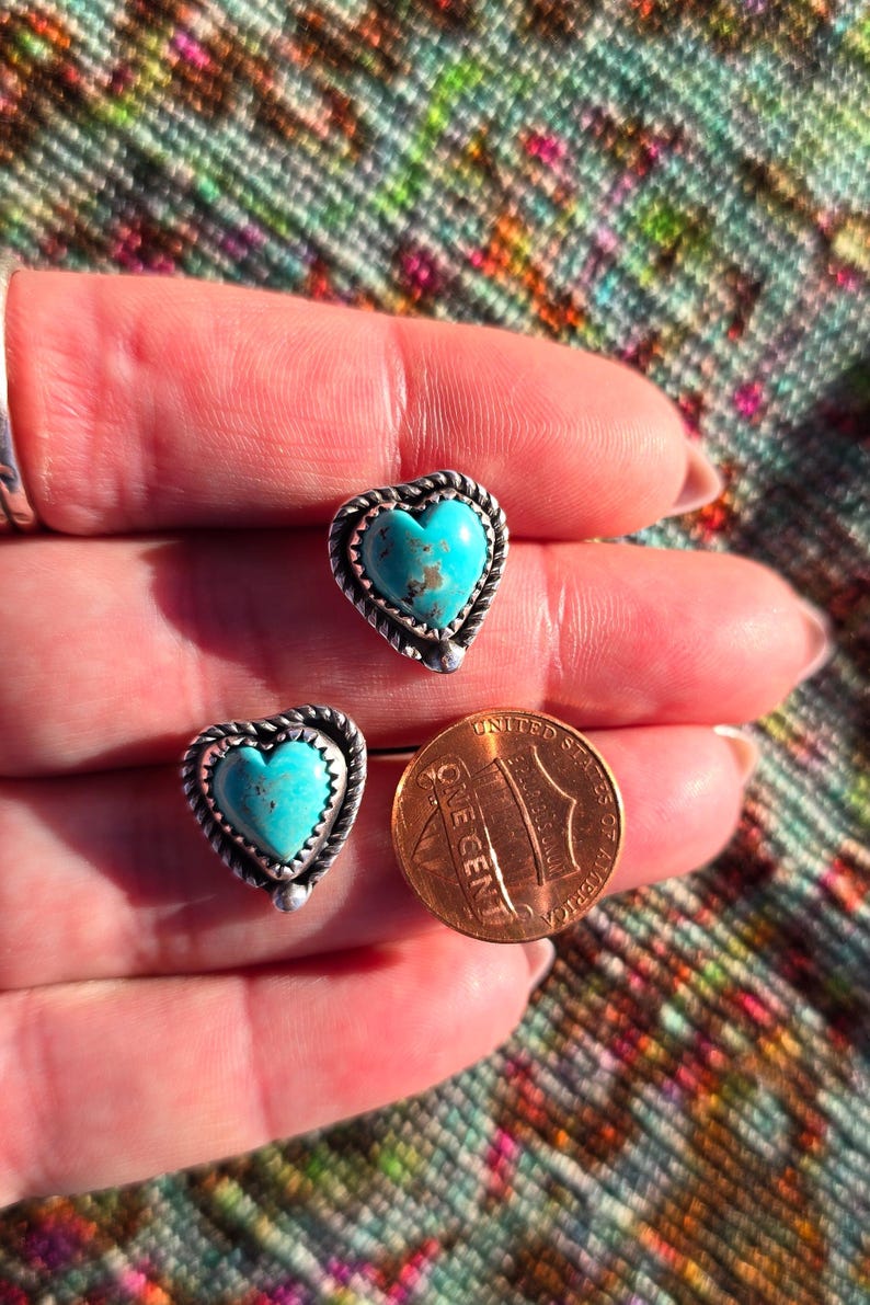 May include: A pair of heart-shaped turquoise stud earrings with a silver rope-like border. A U.S. one-cent coin is next to the earrings for size comparison. The earrings are set against a colorful, patterned background.