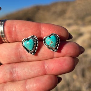 May include: A pair of heart-shaped stud earrings. Each earring features a turquoise stone set in a silver-colored metal frame with a twisted rope design. The earrings are displayed against a blurred outdoor background.