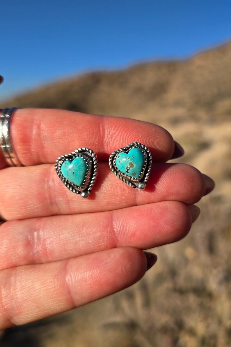 May include: A pair of heart-shaped stud earrings. Each earring features a turquoise stone set in a silver-colored metal frame with a twisted rope design. The earrings are displayed against a blurred outdoor background.