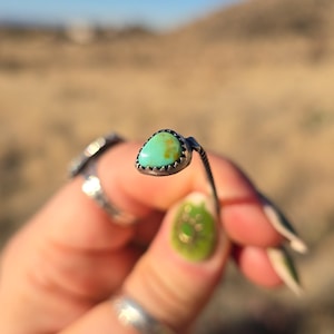 Puede incluir: Anillo de plata con una piedra turquesa en forma de lágrima. El anillo tiene un engaste detallado. El fondo es una escena exterior borrosa.