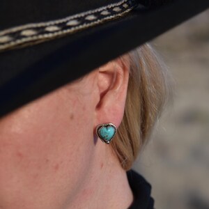 May include: Heart-shaped turquoise stud earring with a silver-colored beaded border. The earring is worn on the ear of a person wearing a black hat and black clothing. The jewelry is a Southwestern style.