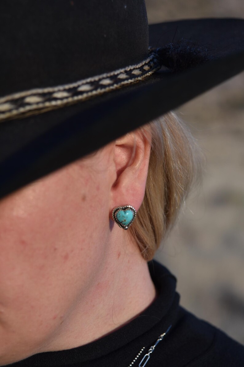May include: Heart-shaped turquoise stud earring with a silver-colored beaded border. The earring is worn on the ear of a person wearing a black hat and black clothing. The jewelry is a Southwestern style.