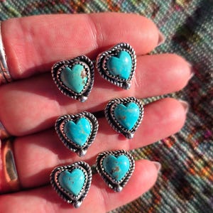 May include: Six silver-toned heart-shaped stud earrings with turquoise centers. Each earring has a twisted rope-like border. The earrings are displayed on a hand against a colorful patterned background.