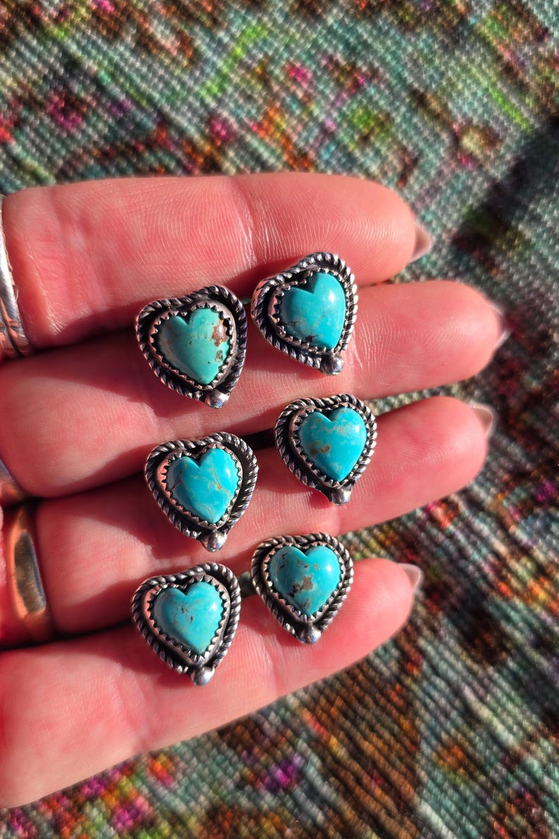 May include: Six silver-toned heart-shaped stud earrings with turquoise centers. Each earring has a twisted rope-like border. The earrings are displayed on a hand against a colorful patterned background.