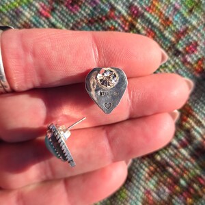 May include: A pair of sterling silver stud earrings. Each earring features a heart-shaped backing with a clear gemstone and the word "STERLING" stamped on it. The earrings have a turquoise stone and a decorative edge.