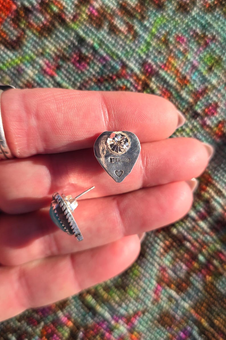 May include: A pair of sterling silver stud earrings. Each earring features a heart-shaped backing with a clear gemstone and the word "STERLING" stamped on it. The earrings have a turquoise stone and a decorative edge.