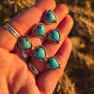 May include: Heart-shaped turquoise stud earrings with a silver rope-like border. The earrings are displayed in a hand, showcasing their vibrant blue-green color and intricate design. The jewelry is perfect for adding a touch of Southwestern style.