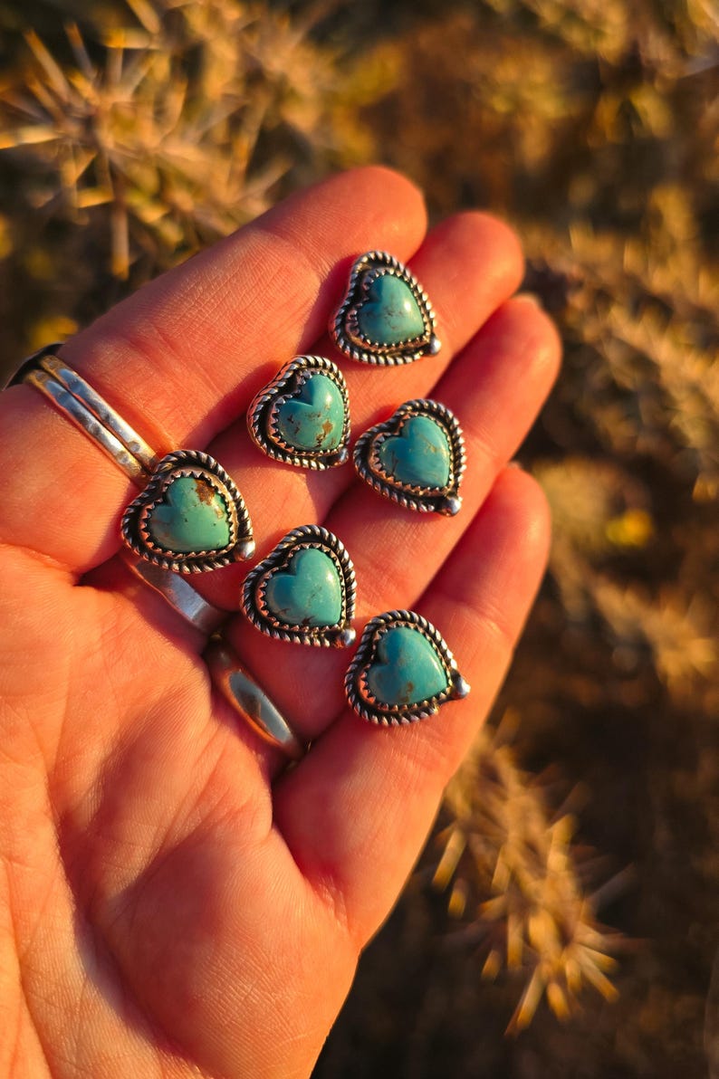 May include: Heart-shaped turquoise stud earrings with a silver rope-like border. The earrings are displayed in a hand, showcasing their vibrant blue-green color and intricate design. The jewelry is perfect for adding a touch of Southwestern style.
