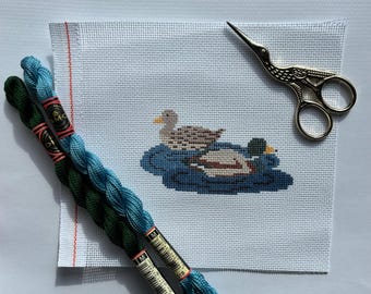 May include: A cross-stitch canvas featuring two ducks on blue water, with green and blue embroidery floss and a decorative bird-shaped scissor. The ducks are brown, white, and yellow, and the water is a dark blue. The floss is in two shades of blue and green.