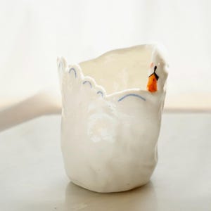 May include: A white ceramic cup with an irregular, hand-molded shape. The rim features a scalloped edge decorated with thin, light blue painted arches. A small, sculpted swan head with an orange beak and black eyes is attached to the rim.