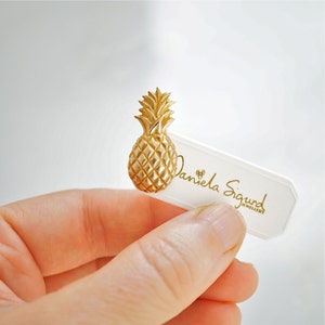 May include: Gold pineapple brooch with a textured surface. The brooch is held in a hand and a white card with the text "Daniela Sigurd Jewellery" is visible in the background.