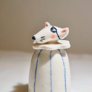 May include: A white ceramic jar with a lid shaped like a rat's head. The rat has a black eye patch and pink accents. The jar is decorated with vertical blue stripes.