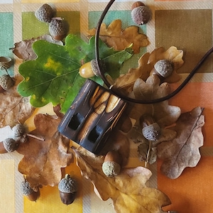 Op de afbeelding: Een handgemaakte houten hangerketting met een donkerbruin leren koord. De rechthoekige hanger heeft gesneden details. De ketting wordt gepresenteerd op een bed van herfstbladeren en eikels, wat een rustiek, door de natuur geïnspireerd thema creëert.