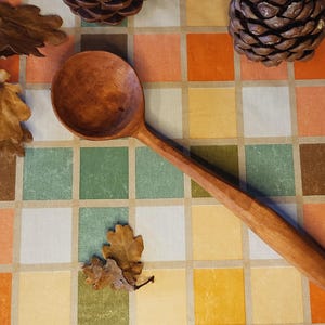 May include: A wooden spoon with a long handle, displayed on a colourful checkered surface. The spoon is a warm brown colour, and the image also includes pine cones and dried leaves.